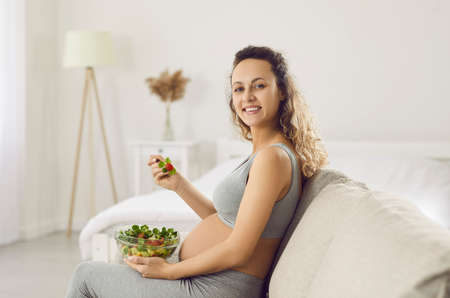 Portrait of smiling young pregnant woman relax at home eat vegetarian salad for dinner. Happy future mother enjoy tasty vegetable lunch rest on maternity leave. Pregnancy and healthcare.の写真素材