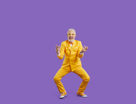 Excited aged man in eccentric suit isolated on purple studio background celebrate win or victory. Smiling mature male in cool bright costume have fun dancing and laughing. Happy maturity.の写真素材