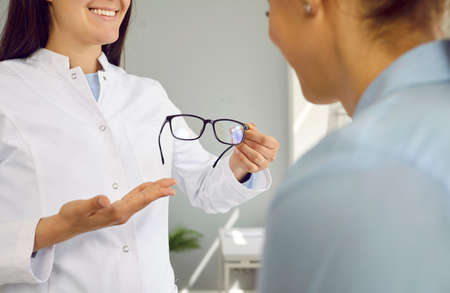 Friendly female optician works with patient and shows her new glasses for testing and trying. Close up of black-rimmed glasses in hands of woman in medical gown. Concept of vision care.の写真素材