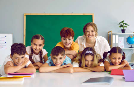 Portrait of cheerful group of elementary school students and their friendly casual female teacher. Joyful schoolchildren leaning on hands laughing at camera with teacher in school classroom.の写真素材