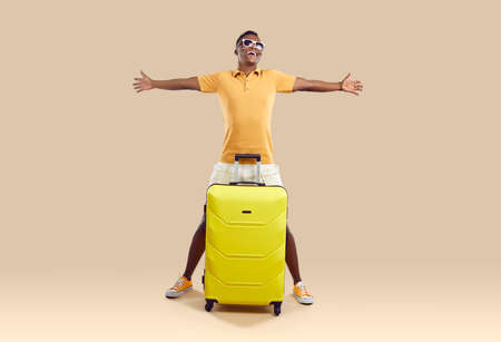 Overjoyed African American man in glasses isolated on brown studio background excited about summer vacation. Smiling biracial guy with suitcase ready for summertime travel or journey.の写真素材