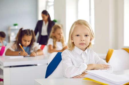 First grader elementary school. Portrait of cute little serious boy sitting at desk during lesson in school classroom. Handsome Caucasian blond kid boy sitting straight and looking at camera.の写真素材