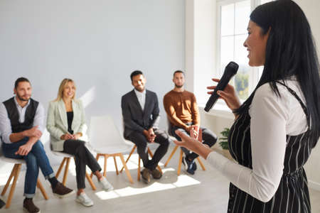 Smiling successful businessman with microphone lead meeting with diverse colleagues. Confident female speaker or coach talk speak at seminar or training with employees in office. Leadership concept.の写真素材