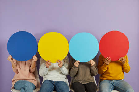 Young people hide their faces behind round colorful blue, yellow and red speech bubble banners that...