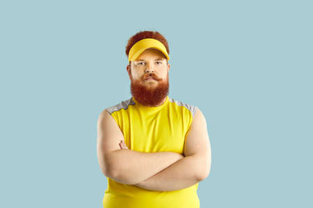 Angry fat man in sportswear. Displeased sports coach standing with his arms crossed and looking at camera. Funny chubby guy with serious unsmiling face expression isolated on blue backgroundの写真素材