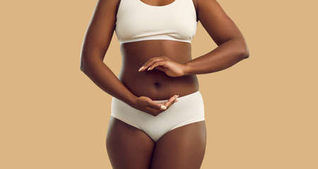 Woman demonstrates gesturing balance in microflora isolated on white background on beige background. Dark skinned plus size model dressed in lingerie holding her hands in circle around her belly.の写真素材