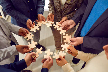 Creative business team finds a solution to the problem. Group of multiracial people form a circle from different pieces of wooden jigsaw puzzle. Teamwork concept. Cropped shot. Conceptual backgroundの写真素材
