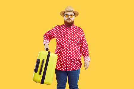 Stylish chubby man with serious expression holding heavy suitcase on orange background. Red-bearded man in straw hat, sunglasses, jeans and red shirt in white peas picks up suitcase looking at camera.の写真素材