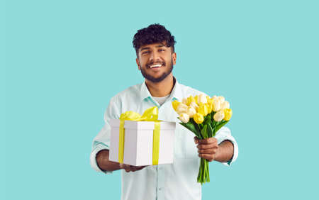 Happy attractive young Indian man millennial holds flowers and gift box for girlfriend smiles...