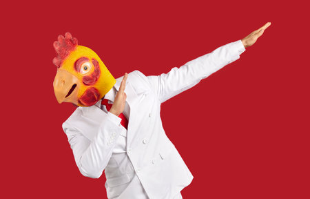 Weirdo man in chicken mask makes movement of famous internet meme about victory on red background. Young man in white suit and with rubber head mask makes dabbing movement during costume party. Bannerの写真素材