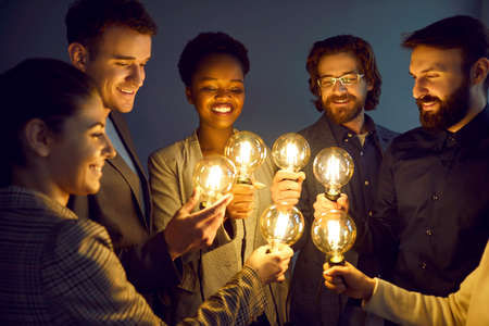 Group of happy smart people hold bright, shining light bulbs as symbol of creative ideas. Smiling multiracial men and women in business attire stand together with light bulbs on in dimly lit office.の写真素材