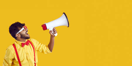 Profile view of funny loud young Indian man in glasses, shirt and red bow tie isolated on solid yellow colour copy space banner background shouting through megaphone and making big sale announcementの写真素材