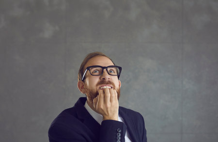 Nervous doubtful adult funny businessman biting finger nail wondering, freaking out and looking up on copy space standing against grey studio background. Human emotion expression. Closeup shotの写真素材