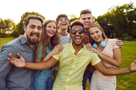 Group of young diverse people enjoy free time in summer, meet up in the park and have fun all together. Bunch of six happy excited multi ethnic friends looking at the camera, smiling and laughingの写真素材