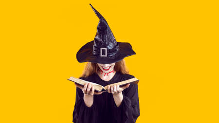 Woman in Halloween costume looking through book of spells. Cunning evil witch with bloody neck cut and creepy happy smile hides face under hat and chooses most terrible curse from witchcraft manualの写真素材