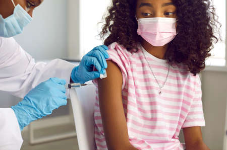 Woman who works as doctor or nurse gives arm injection to little child. Afro American school girl in facemask gets flu, covid or monkeypox vaccine shot to develop strong immunity to dangerous virusの写真素材