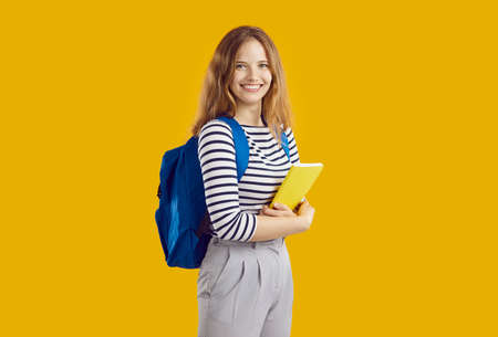 Portrait of joyful female student with backpack and textbook on orange background. Beautiful teenage...