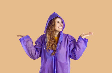 Woman with long wavy blond hair posing in purple raincoat isolated on beige background. Happy young girl enjoying rainy weather and holding palms facing up to catch falling raindrops. Fashion conceptの写真素材