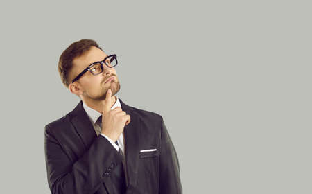 Businessman thinking about something. Young business man in suit and glasses standing isolated on gray background, holding hand on chin and looking at copy space with pensive puzzled face expressionの写真素材