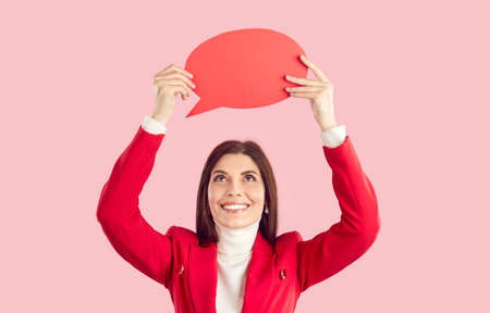 Pretty lady sharing opinion and showing message bubble. Happy cheerful beautiful young woman in red jacket standing on pink studio background, holding up red empty mock up speech balloon and smilingの写真素材
