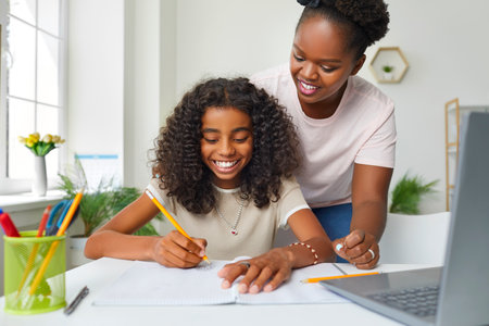 Help with homework. Joyful african american teenage girl doing school homework at home with moms help. Smiling young dark-skinned woman watches her daughter write in notebook. Education and family.の写真素材