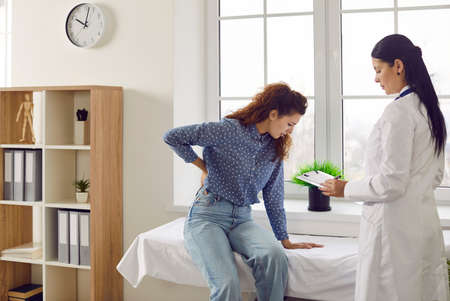 Woman complains to doctor about back or kidney pain during medical examination in hospital room. Female doctor records complaints of patient who is holding her back while sitting on medical couch.の写真素材