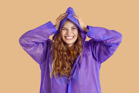 Happy woman in a raincoat having fun in rainy weather. Cheerful excited young woman or a teen girl in a purple rain coat standing isolated on a beige background, looking at the camera and smilingの写真素材