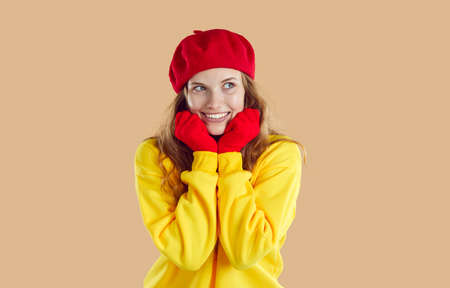 Happy smiling woman with dreamy facial expression isolated on light beige background. Beautiful Caucasian woman in warm casual sweatshirt, red beret and gloves is flirtatiously looking to the side.の写真素材