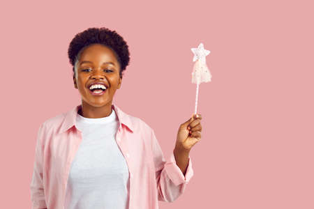 Portrait of happy funny modern fairy. Studio shot of joyful positive woman with magic wand. Young African American girl in casual shirt standing isolated on pink background, holding wand and smilingの写真素材