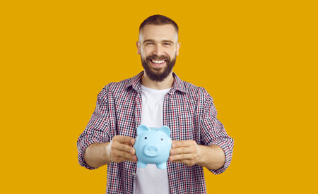 Joyful young casual Caucasian man holding piggy bank for coins and cash holding it out to you as invitation to personal financial planning standing in orange studio. Economics and saving moneyの写真素材