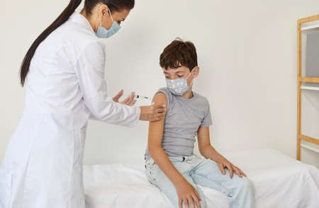 Child getting vaccine injection for covid, flu, monkeypox or other infectious disease. Nurse or doctor giving shot to school kid who sitting on couch in medical office. Childrens vaccination conceptの写真素材