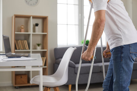 Cropped indoor shot of weak young man with disability or plaster cast bandage on broken injured leg using axillary underarm crutches and walking to working desk with laptop computer in home workplaceの写真素材