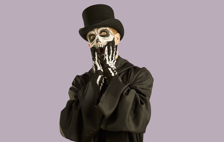 Portrait of shocked man in Halloween make-up and costume grabbing his face in fright. Man in black hat, suit and skull make-up opens his eyes and mouth wide in fear on light lilac background.の写真素材