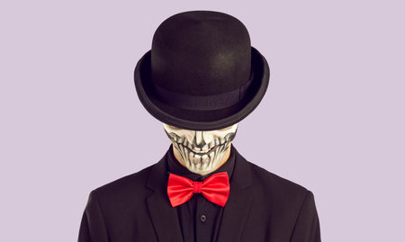 Studio shot of skeleton man in stylish Halloween outfit. Portrait of man with skull makeup dressed in black suit and red bow tie hiding his face under black hat isolated on light purple backgroundの写真素材
