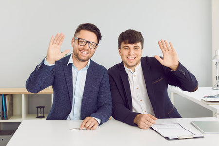 Webcam portrait of happy business people starting work video call. Two young men in suits sitting at office desk, looking at camera, smiling, waving hello, greeting colleagues at online conferenceの写真素材