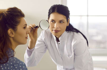 Female doctor looking at patients skin through magnifying glass. Professional dermatologist and skincare specialist investigating moles or tumor growth signs on womans face. Skin checkup conceptの写真素材