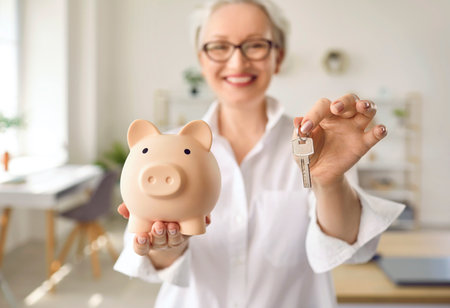 Happy smiling middle aged woman holding a pink piggy bank and house keys. Soft focus, close up. Concept of saving money, taking mortgage loan, buying new houseの写真素材