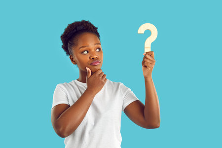 Young dark skinned woman with doubtful expression is looking at wooden question mark in her hand on...