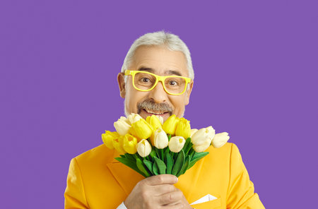 Portrait of cheerful cute old man with bouquet of yellow tulips on purple background. Positive senior man in stylish bright clothes with funny expression looks at camera holding flowers. Close up.の写真素材