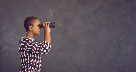 Banner web image of millennial African American woman on black studio background hold binocular glass look at empty mockup copy space. Happy young ethnic girl use optical equipment consider deal.の写真素材