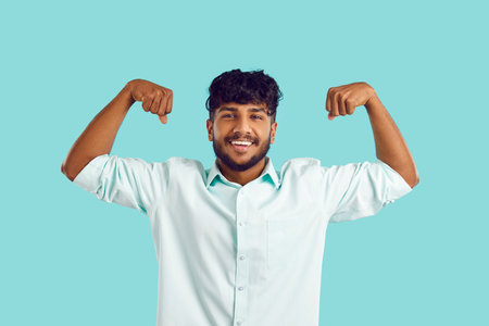 Portrait of smiling Indian man isolated on blue studio background show muscles feel powerful. Happy...