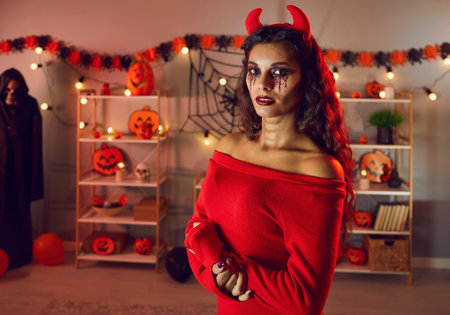 Portrait of woman in evil costume with horns, scary dark makeup celebrating Halloween indoors. Young female on ball masquerade on All Saints Hallows eve in decorated home interior. Festive.の写真素材