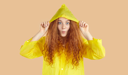 Cheerful emotional young woman in raincoat looking at camera with funny expression. Portrait of red-haired girl with wavy hair wearing yellow raincoat with hood on light beige background in studio.の写真素材