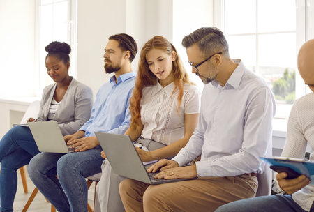 Diverse people using laptop computers while sitting in line in office workplace. Group of multiracial young and mature men and women sitting in row, using notebook PCs and working on business projectsの写真素材