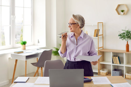 Senior business lady in office uses voice recognition function or virtual assistant on mobile phone. Friendly mature entrepreneur answers an incoming call using speakerphone or records voice message.の写真素材