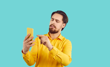 Focused young man look at cellphone screen frustrated by news online. Pensive guy browse wireless internet on smartphone gadget. New technologies and communication. Blue studio background.の写真素材