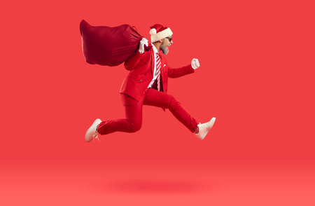 Happy excited man in Santa Claus hat and red modern suit running on red studio background, carrying big sack with presents and jumping high in air. Christmas sale and present delivery conceptの写真素材