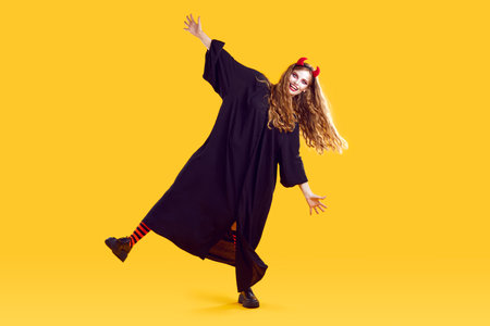 Full length funky funny woman in halloween costumewith terrifying makeup, red devil horns is dancing on yellow background. Halloween celebration, costume party, masquerade, fear and horror concept.の写真素材