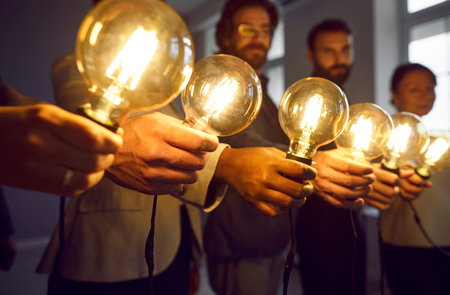Glowing light bulbs in hands of creative business team in office as symbols of their ideas. Close up of turned on Edison bulbs in hands of men and women standing in row. Selected focus.の写真素材