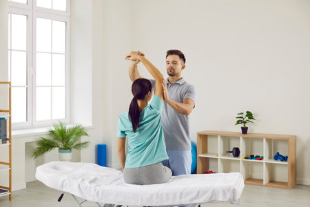Doctor at modern physical therapy and rehabilitation clinic examining young womans arm. Male...
